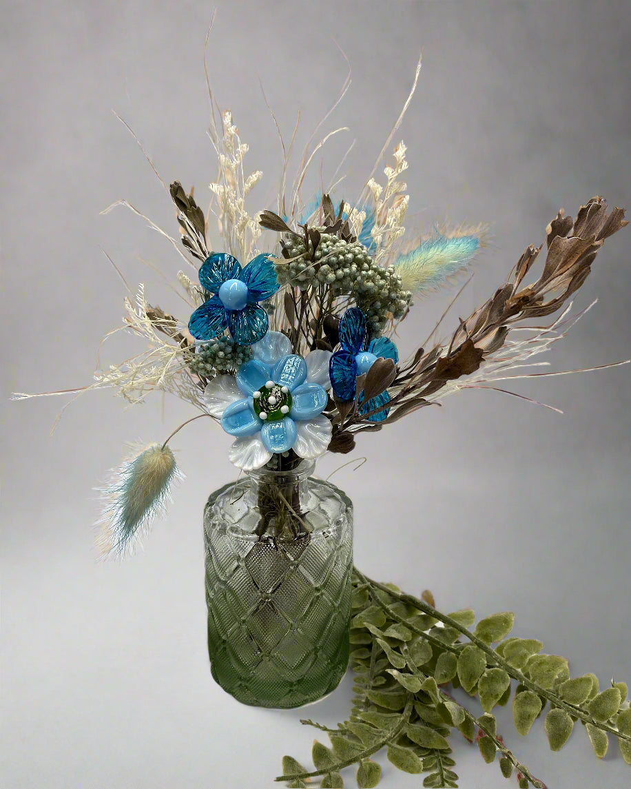 Decorative floral arrangement with blue flowers and green leaves in a textured glass vase on a light background.